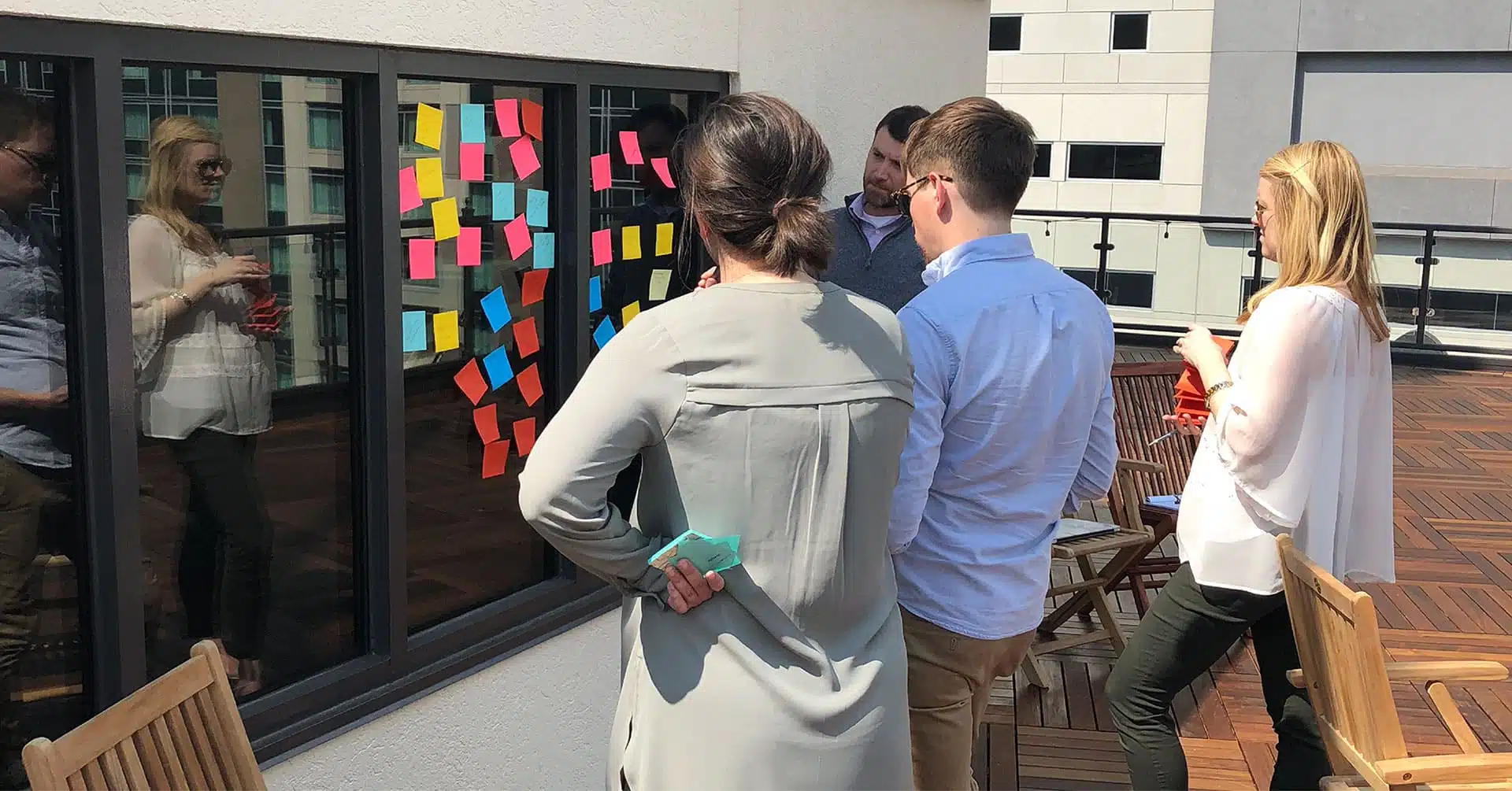 A group of people standing on an outdoor rooftop terrace participating in a brainstorming session with colorful sticky notes on a window.