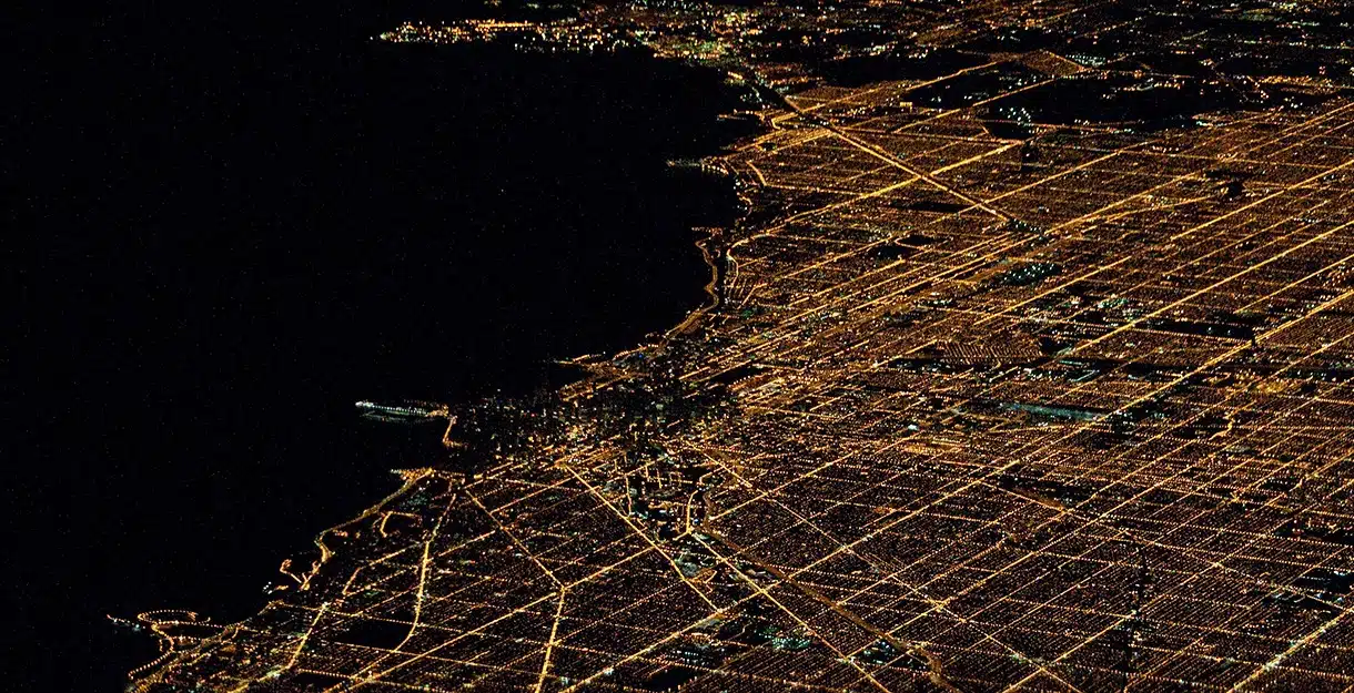 BGM21 321750 GridPerspectives An aerial nighttime view of a brightly lit city coastline with a grid of golden lights bordering a dark body of water.