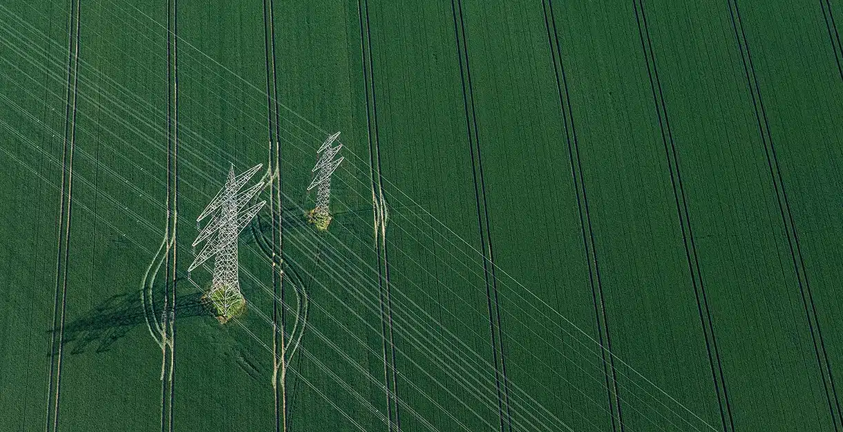 An aerial view of high-voltage power lines and transmission towers standing in a vast, vibrant green agricultural field.