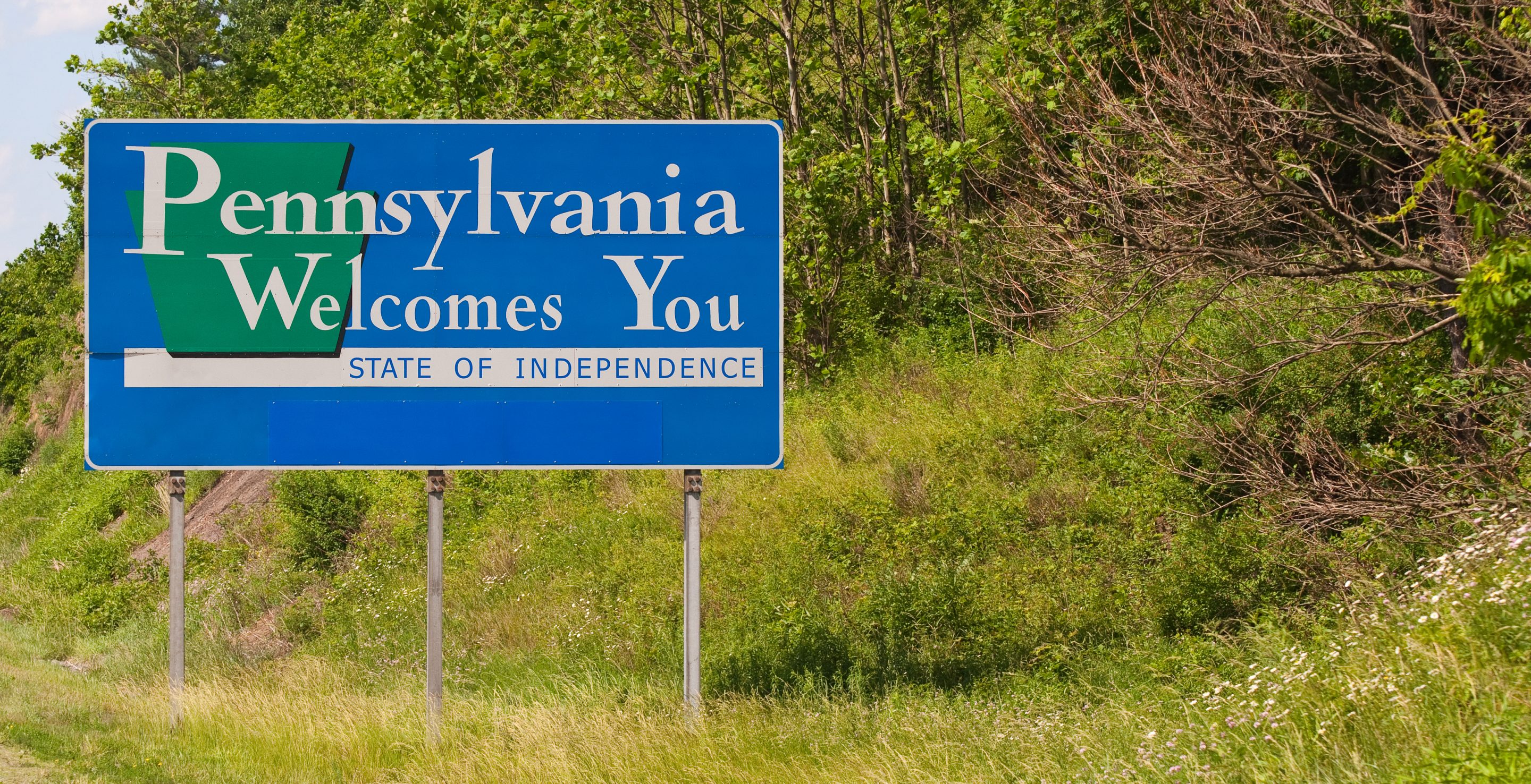 A large blue roadside sign that reads "Pennsylvania Welcomes You" with the slogan "State of Independence" against a backdrop of green trees.