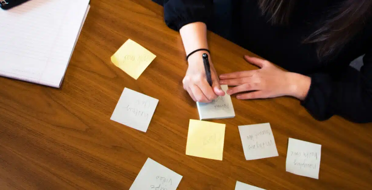 A person writing on a sticky note at a wooden desk surrounded by other scattered notes.