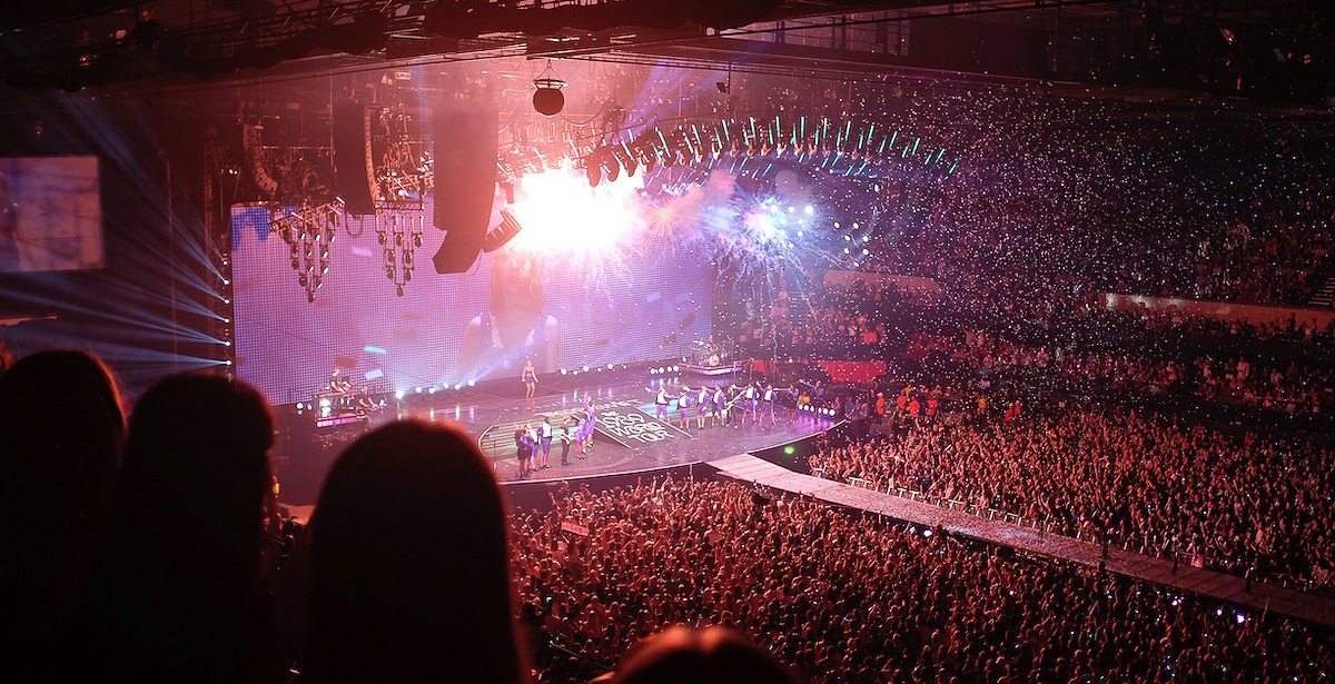 Taylor Swift on tour A wide shot of a massive indoor stadium concert during Taylor Swift's "1989 World Tour," featuring a crowded arena, a glowing stage, and confetti falling from the ceiling.