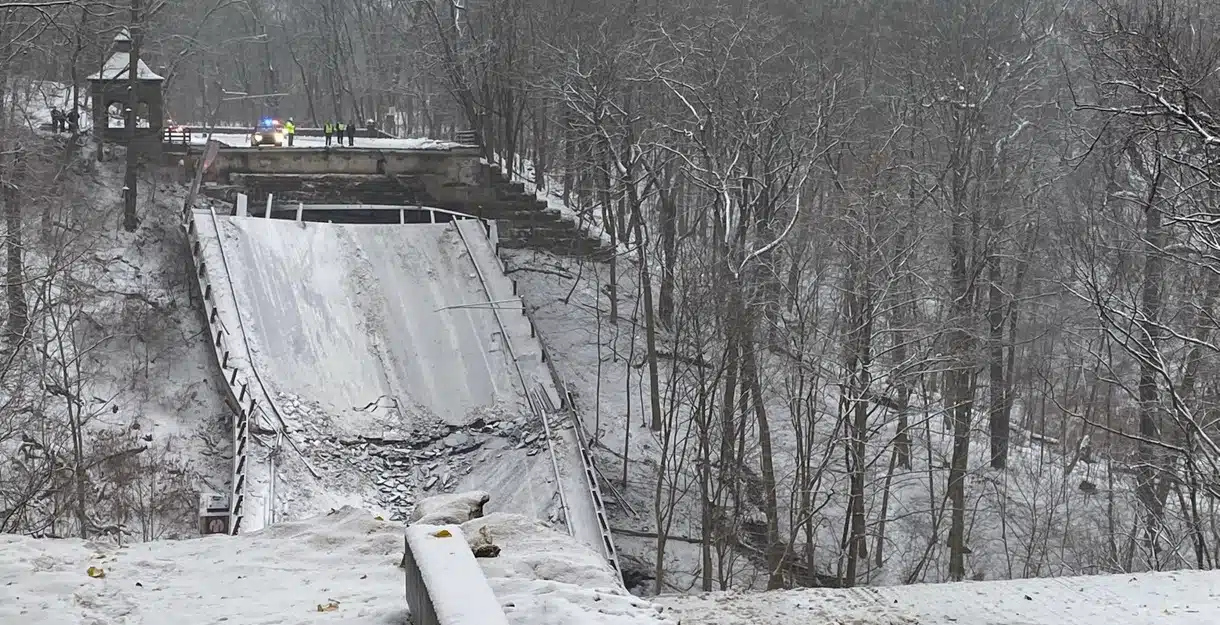 A snowy winter scene showing the dramatic structural collapse of the Fern Hollow Bridge in Pittsburgh, with the bridge deck buckled into the ravine below.
